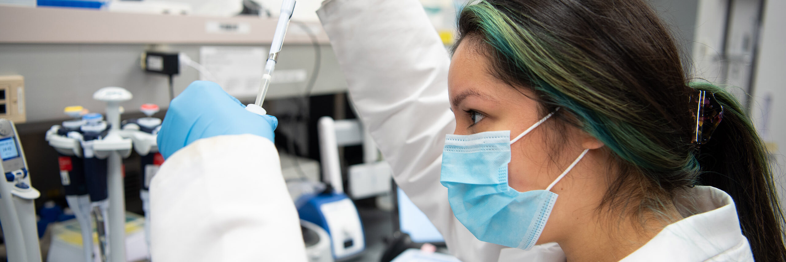 A scientist in a lab coat and mask holds a tube, conducting experiments in a laboratory setting.