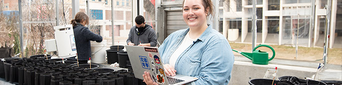 Woman sitting in a greenhouse, holding a laptop.